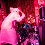 A photo bathed in red light shows the duo Radioactive Vampire Teeth performing on the stage upstairs at The Palomino. The drummer is seen performing the background, and the frontperson looks to be smashing the symbals from the front. 