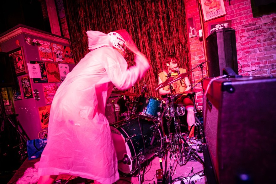 A photo bathed in red light shows the duo Radioactive Vampire Teeth performing on the stage upstairs at The Palomino. The drummer is seen performing the background, and the frontperson looks to be smashing the symbals from the front. 