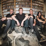 Four members of the group Salthorse are seen sitting on the floor against a wooden shelf holding CDs. They are sitting on the floor with a black and white abstract pattern on it. 