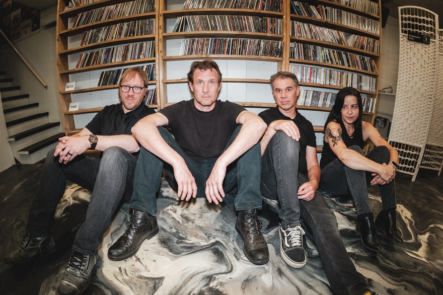 Four members of the group Salthorse are seen sitting on the floor against a wooden shelf holding CDs. They are sitting on the floor with a black and white abstract pattern on it. 