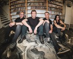 Four members of the group Salthorse are seen sitting on the floor against a wooden shelf holding CDs. They are sitting on the floor with a black and white abstract pattern on it. 