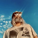 A colour photo taken from below shows artist Ryan Bourne standing outside with a bright blue sky behind them. They are are wearing shimmering sunglasses, a tan t-shirt and a tan ballcap.