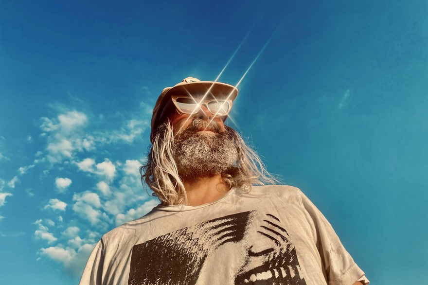 A colour photo taken from below shows artist Ryan Bourne standing outside with a bright blue sky behind them. They are are wearing shimmering sunglasses, a tan t-shirt and a tan ballcap.