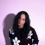 A colour photo shows the frontperson of the band sadbirthdays posing in front of a light purple background. They are seen wearing a black knit sweater with daisies on it. 