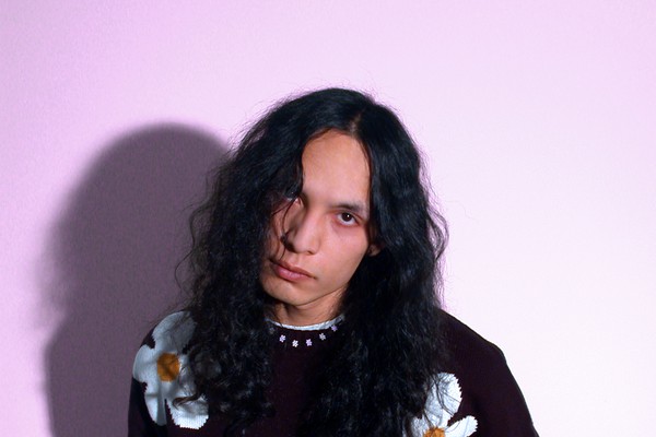 A colour photo shows the frontperson of the band sadbirthdays posing in front of a light purple background. They are seen wearing a black knit sweater with daisies on it. 