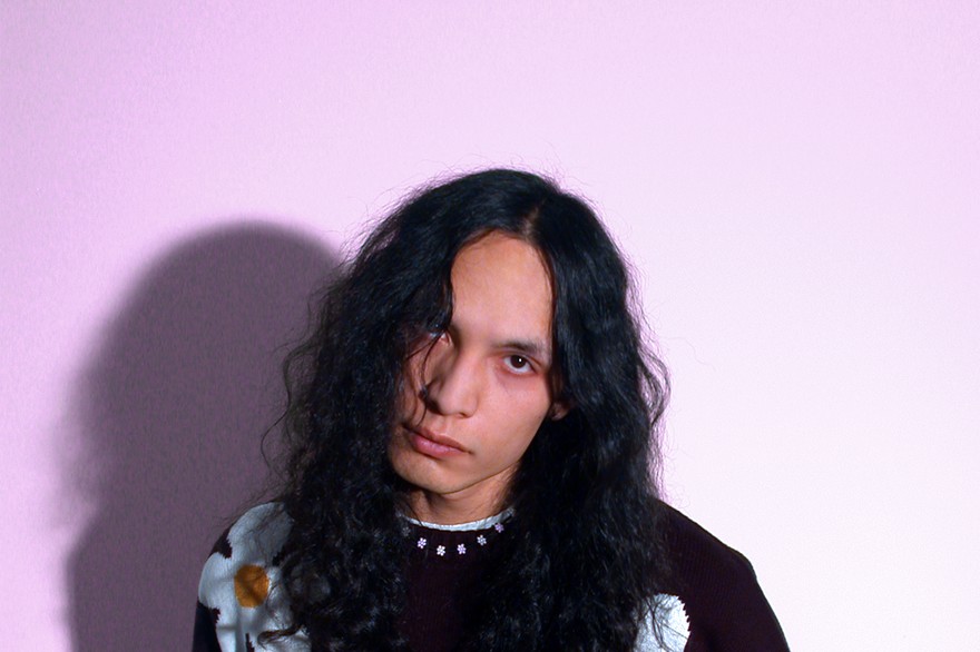 A colour photo shows the frontperson of the band sadbirthdays posing in front of a light purple background. They are seen wearing a black knit sweater with daisies on it. 