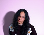 A colour photo shows the frontperson of the band sadbirthdays posing in front of a light purple background. They are seen wearing a black knit sweater with daisies on it. 