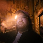Artist SAMWOY is shown in a close-up photo standing in a room with wooden walls covered in graffiti. A sole lightbulb in the background lights the room. 