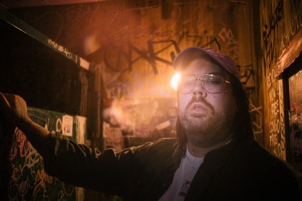 Artist SAMWOY is shown in a close-up photo standing in a room with wooden walls covered in graffiti. A sole lightbulb in the background lights the room. 