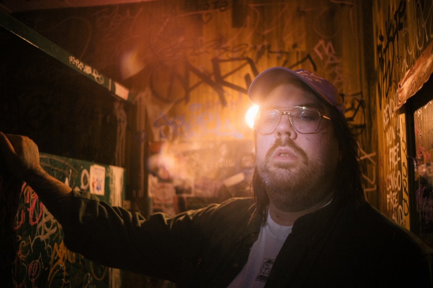 Artist SAMWOY is shown in a close-up photo standing in a room with wooden walls covered in graffiti. A sole lightbulb in the background lights the room. 