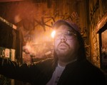 Artist SAMWOY is shown in a close-up photo standing in a room with wooden walls covered in graffiti. A sole lightbulb in the background lights the room. 