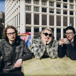 A colour photo shows three-piece band schoolyard sitting outdoors at picninc table in an inner city location with several large building seen in the background.