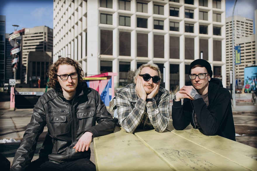 A colour photo shows three-piece band schoolyard sitting outdoors at picninc table in an inner city location with several large building seen in the background.