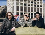 A colour photo shows three-piece band schoolyard sitting outdoors at picninc table in an inner city location with several large building seen in the background.