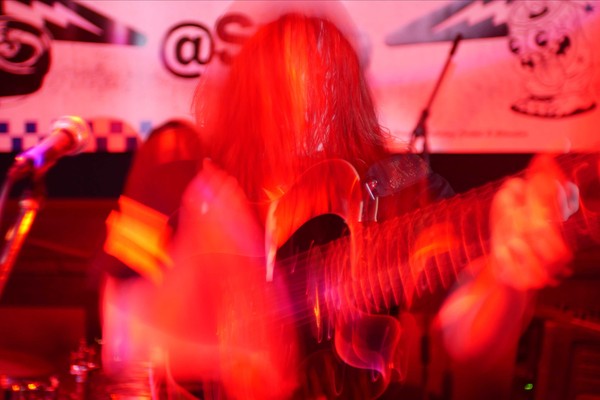 A hazy colour photo bathed in red light shows a member of the band SENTRIES playing a guitar. A long exposure effect distorts the photo.