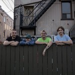 A colour photo shows four-piece band Sea of Lettuce standing behind a brown fence with their arms folded on top. Several older buildings are shown in the background. 