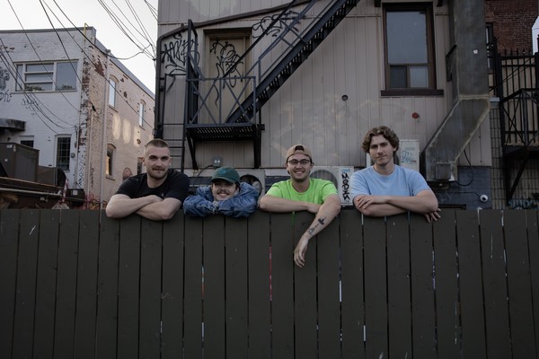 A colour photo shows four-piece band Sea of Lettuce standing behind a brown fence with their arms folded on top. Several older buildings are shown in the background. 