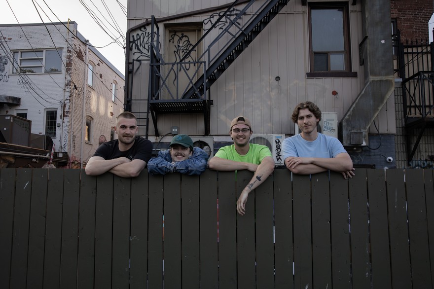 A colour photo shows four-piece band Sea of Lettuce standing behind a brown fence with their arms folded on top. Several older buildings are shown in the background. 