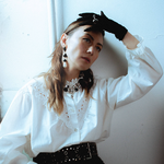 A colour photo shows artist Sheenah Ko sitting near in a window in a white room posing with their left hand touching their forehead. The are wearing black gloves, a white blouse and a brown dress.