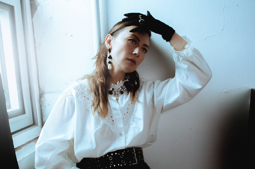 A colour photo shows artist Sheenah Ko sitting near in a window in a white room posing with their left hand touching their forehead. The are wearing black gloves, a white blouse and a brown dress.