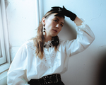 A colour photo shows artist Sheenah Ko sitting near in a window in a white room posing with their left hand touching their forehead. The are wearing black gloves, a white blouse and a brown dress.