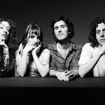 A black and white photo shows four-piece band Shunk posing against a black background, resting their arms on a black platform placed in front of them. 