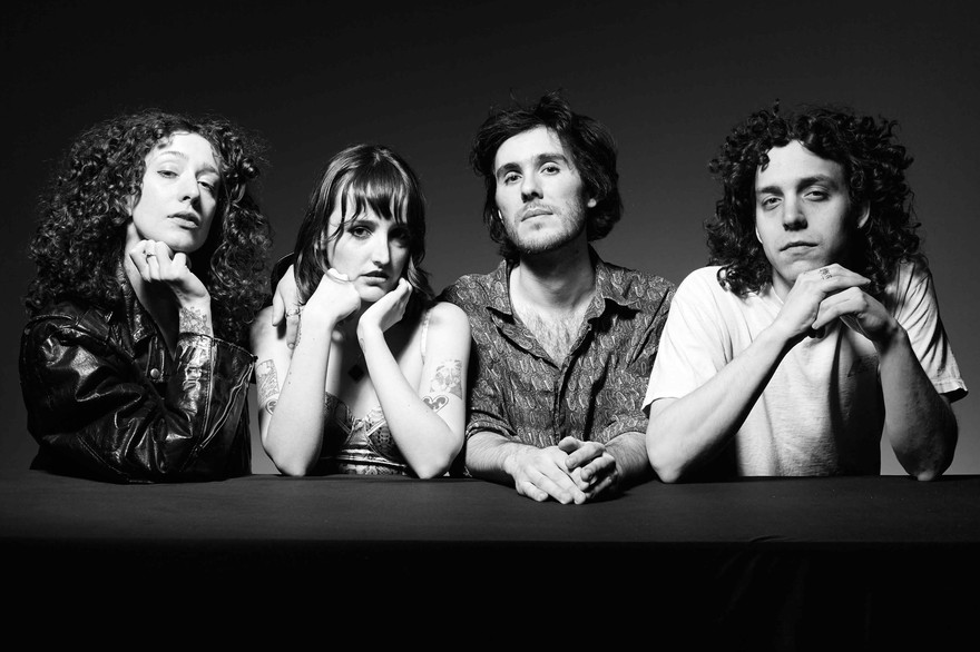 A black and white photo shows four-piece band Shunk posing against a black background, resting their arms on a black platform placed in front of them. 