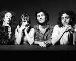 A black and white photo shows four-piece band Shunk posing against a black background, resting their arms on a black platform placed in front of them. 