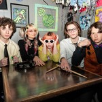 A colour photo shows five-piece group SHY FRiEND sitting at a square table looking at the camera. A tambourine and drum sticks sit on top, and colourful artwork is hanging on the grey wall behind them. 
