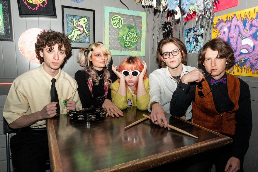 A colour photo shows five-piece group SHY FRiEND sitting at a square table looking at the camera. A tambourine and drum sticks sit on top, and colourful artwork is hanging on the grey wall behind them. 