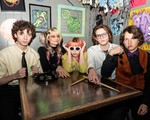 A colour photo shows five-piece group SHY FRiEND sitting at a square table looking at the camera. A tambourine and drum sticks sit on top, and colourful artwork is hanging on the grey wall behind them. 