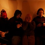 A colour photo shows four-piece group Sloosh standing in a line in a dimly lit room. Two members look down at their gameboys, one is covering their face with their hands, and the member on the left is wearing a knit red devil mask. 