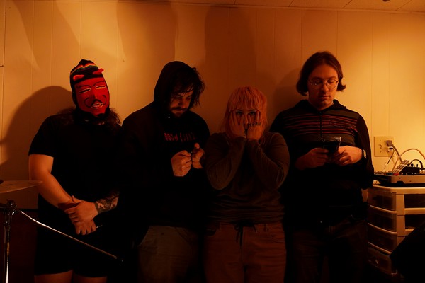 A colour photo shows four-piece group Sloosh standing in a line in a dimly lit room. Two members look down at their gameboys, one is covering their face with their hands, and the member on the left is wearing a knit red devil mask. 