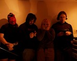A colour photo shows four-piece group Sloosh standing in a line in a dimly lit room. Two members look down at their gameboys, one is covering their face with their hands, and the member on the left is wearing a knit red devil mask. 