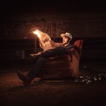 A colour photo shows the frontperson of the group Spank Williams sitting outside in an old reclining chair reading a flaming newspaper with a pile of empty cans on the ground next to them. 