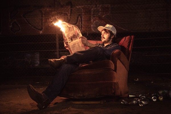A colour photo shows the frontperson of the group Spank Williams sitting outside in an old reclining chair reading a flaming newspaper with a pile of empty cans on the ground next to them. 