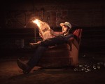 A colour photo shows the frontperson of the group Spank Williams sitting outside in an old reclining chair reading a flaming newspaper with a pile of empty cans on the ground next to them. 