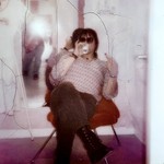 A cracked fuzzy photo shows a member of the band Spirit Hotel sitting in a red chair, drinking from a can and wearing sunglasses holding up a middle finger at the camera. A mirror on the wall behind them shows the photographer taking the photo with the flash going off. 