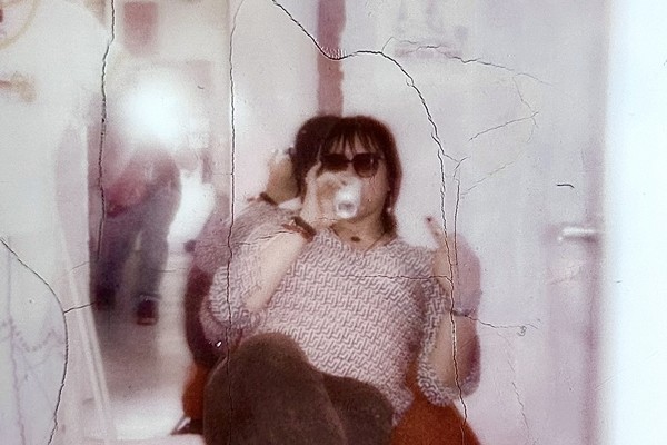 A cracked fuzzy photo shows a member of the band Spirit Hotel sitting in a red chair, drinking from a can and wearing sunglasses holding up a middle finger at the camera. A mirror on the wall behind them shows the photographer taking the photo with the flash going off. 