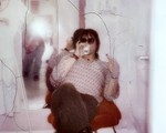 A cracked fuzzy photo shows a member of the band Spirit Hotel sitting in a red chair, drinking from a can and wearing sunglasses holding up a middle finger at the camera. A mirror on the wall behind them shows the photographer taking the photo with the flash going off. 