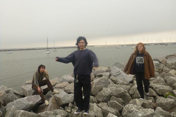 Three members of the group Still Depths are seen standing on rocks with a body of water behind them. Many sailboats are shown on the water in the background. 