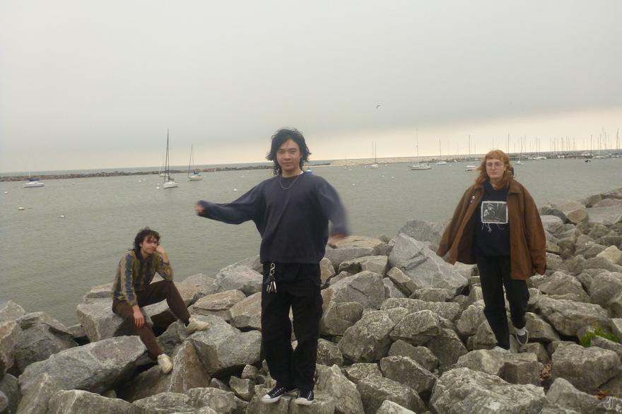 Three members of the group Still Depths are seen standing on rocks with a body of water behind them. Many sailboats are shown on the water in the background. 