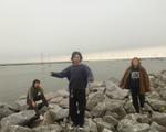 Three members of the group Still Depths are seen standing on rocks with a body of water behind them. Many sailboats are shown on the water in the background. 