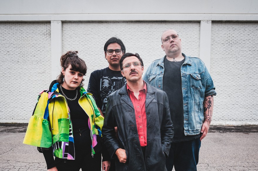A colour photo of four-piece band Stucco shows them standing on a cracked concrete ground with a white brick building in the background. 