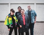 A colour photo of four-piece band Stucco shows them standing on a cracked concrete ground with a white brick building in the background. 