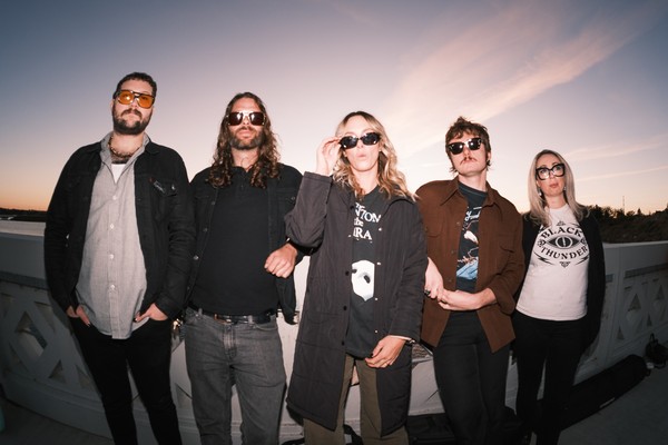 A slightly blurry photo shows five-piece band Sub / / Cell standing on a white bridge with each member wearing sunglasses. A blue and light purple sky is shown behind them. 