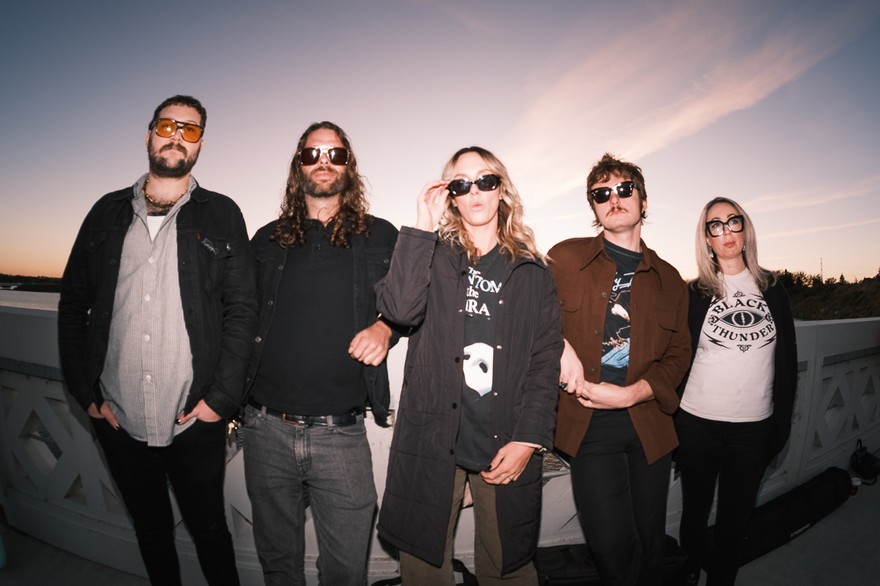 A slightly blurry photo shows five-piece band Sub / / Cell standing on a white bridge with each member wearing sunglasses. A blue and light purple sky is shown behind them. 