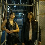 A colour photo shows duo Sunflower Medicine standing in an alley under scaffolding and next to a chainlink maintenance closet. 