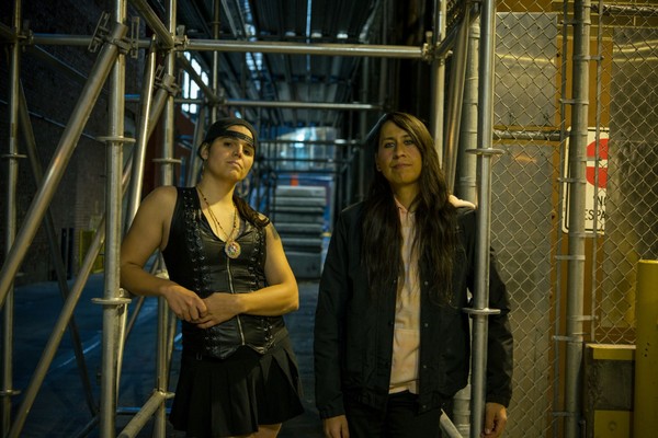 A colour photo shows duo Sunflower Medicine standing in an alley under scaffolding and next to a chainlink maintenance closet. 