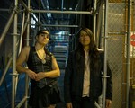 A colour photo shows duo Sunflower Medicine standing in an alley under scaffolding and next to a chainlink maintenance closet. 
